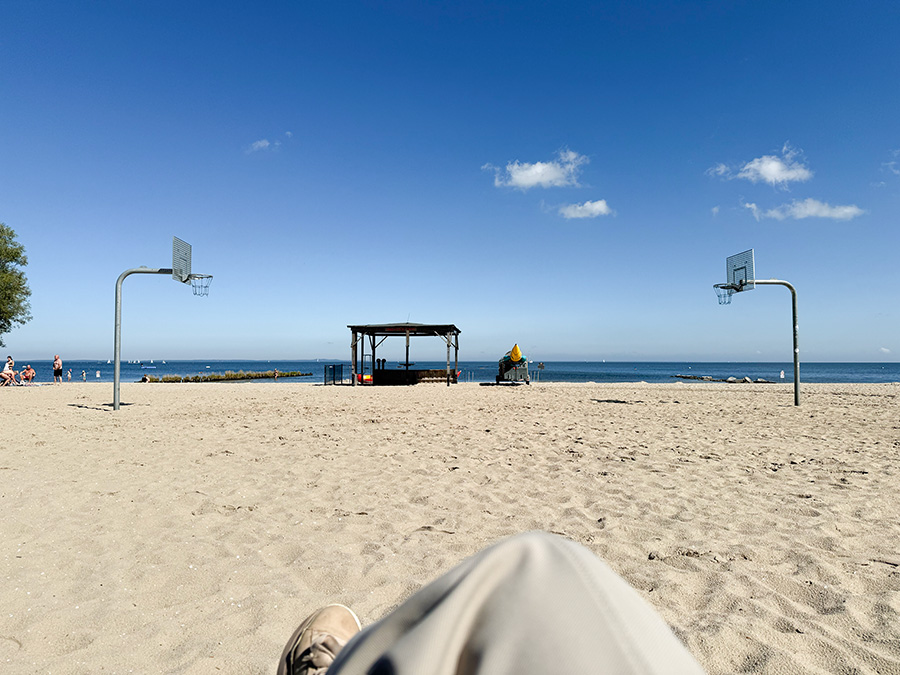 Strand, Sand, Sonne, Basketballkorb, blauer Himmel, beige Hose, beige Sneakers